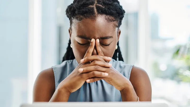 African woman suffering with a headache and feeling stressed about deadlines while working on a laptop in an office alone