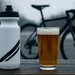Bidon en pint bier naast elkaar op een houten tafel, met een racefiets onscherp op de achtergrond in een grijze winterse setting.