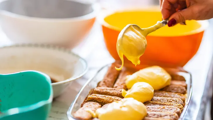 Close up of female hand putting and spreading mascarpone cream with spoon on tiramisu. Homemade preparation of italian cake tiramisu. Selective focus. Blurred terrines and bowls in the background