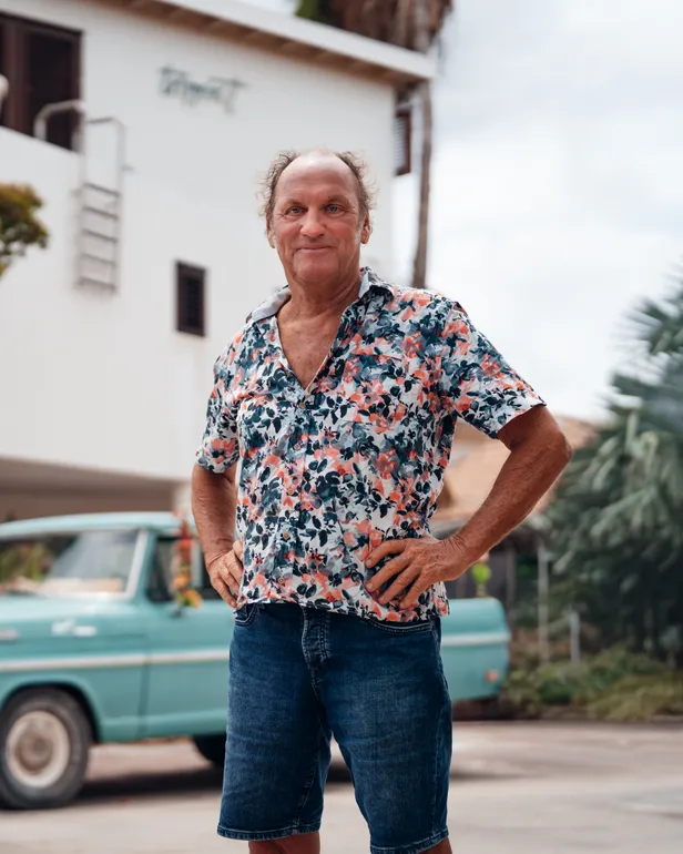 Fred poseert lachend voor zijn B&B en blauwe auto op het zonnige Bonaire voor B&B Vol Liefde 2026. 