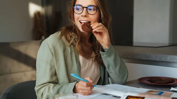 Pleased nice student woman eating cookie while doing homework at home