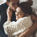 Caucasian man kissing his cute wife dressed in a white knitted sweater embracing in bed