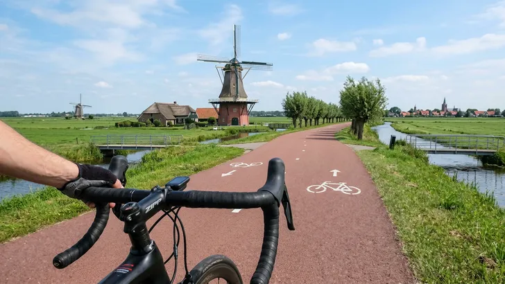 Een POV-foto vanaf een racefiets waarbij de wielrenner met een gehandschoende hand de shifter bedient op een vlakke weg, met een naderende steile brug in de verte in een zonnig Nederlands landschap.