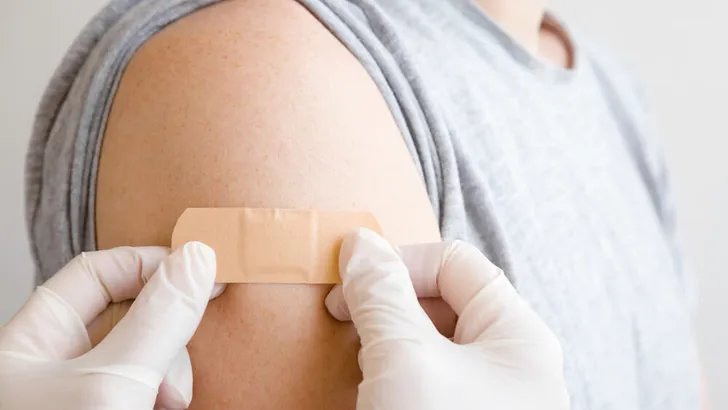 Doctor in white rubber protective gloves putting adhesive bandage on young man's arm after scratch on skin or injection of vaccine. First aid. Medical, pharmacy and healthcare concept. Closeup.