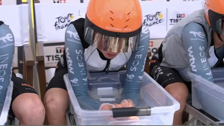 Drie wielrenners van de INEOS Grenadiers wielerploeg zitten naast elkaar op stoelen met hun armen diep in transparante bakken met ijswater om hun lichaam te koelen voor de start van de ploegentijdrit in de Tour de France.