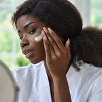 Attractive black young woman applying cream on face skin looking in mirror.