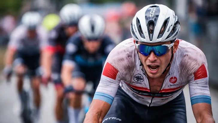 Vermoeide wielrenner raakt het wiel kwijt in het peloton tijdens de koers, gezicht vertrokken van inspanning in het tweede uur.