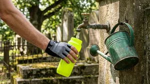 Een close-up van een felgekleurde, moderne wielerbidon die wordt gevuld met water onder een oud, koperen kraantje op een begraafplaats. Op de zonnige en onscherpe achtergrond zijn de contouren van grafstenen en groene struiken zichtbaar.