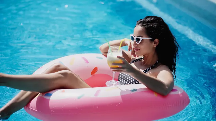 cheerful woman in sunglasses holding glass with cocktail and swimming on inflatable ring in pool