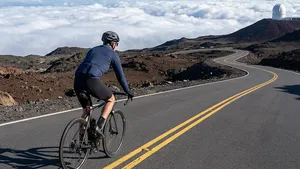 Mannelijke wielrenner met helm beklimt een vulkaan op Hawaï en fietst over een geasfalteerde weg boven een indrukwekkend wit wolkendek.