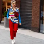 Fashionable Woman wearing trendy outfit, red trousers, blue jacket, striped vest, white shoes and yellow sunglasses. Model with short hair walking the street