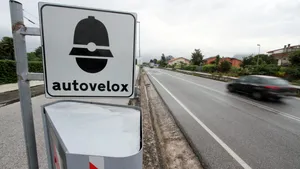 Italiaans autovelox-bord en flitspaal langs een natte weg, met een voorbijrijdende vrachtwagen op de achtergrond.