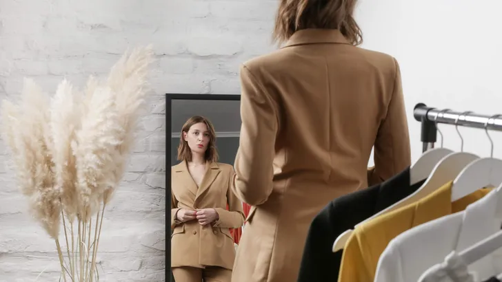 Young woman choosing clothes and trying on beige blazer in fashion atelier or personal wardrobe.