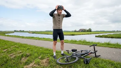Een radeloos kijkende wielrenner met helm en armstukken staat langs een Nederlands fietspad, zuchtend met zijn handen in het haar. Zijn racefiets ligt in de berm met een volledig leeggelopen achterband, in een weids polderlandschap met kanalen en velden.