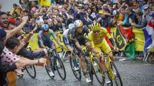 Gele trui Tadej Pogacar leidt het peloton door een natte bocht terwijl fans langs de kant luid juichen tijdens een Tour-etappe.