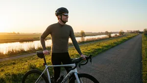 Een mannelijke wielrenner met helm kijkt bij zonsopkomst hoopvol uit over een rustige polderweg, staand naast zijn racefiets.