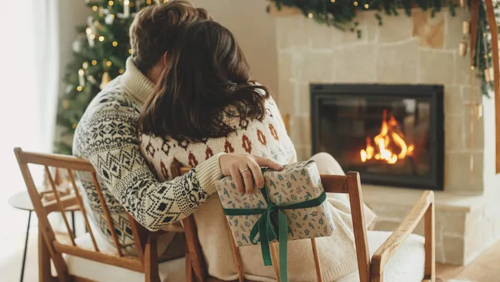 Happy young family in cozy sweaters exchanging stylish christmas gifts on background of fireplace with modern festive mantle and christmas tree with lights. Happy Holidays!