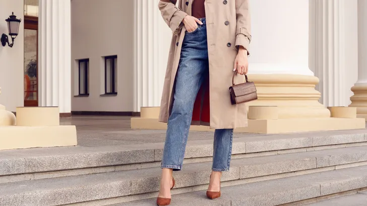 Streetstyle-foto van stijlvolle vrouw in spijkerbroek, pumps en beige trenchcoat.