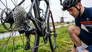 Een mannelijke wielrenner met helm kijkt in een Nederlands polderlandschap naar de achterderailleur van zijn racefiets
