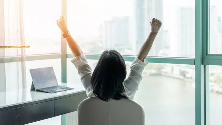 Business achievement concept with happy businesswoman relaxing in office or hotel room, resting and raising fists with ambition looking forward to city building urban scene through glass window