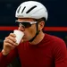 Wielrenner in rode jersey met witte helm en zonnebril drinkt koffie uit een papieren beker bij de teambus.