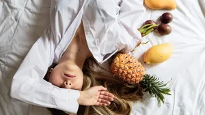 Young woman with long blonde hair wearing white home shirt lying on the bed. Summer healthy raw exotic fruits in bed. Top view, overhead. Time for relax and dream. Healthy eating concept
