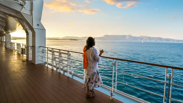 A woman sips a drink on the deck of a cruise ship as the sun sets and the ship passes islands on the Aegean Sea