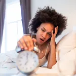 Sleepy young woman stretching hand to ringing alarm to turn it off. Early wake up, not getting enough sleep, getting work concept. waking up with headache