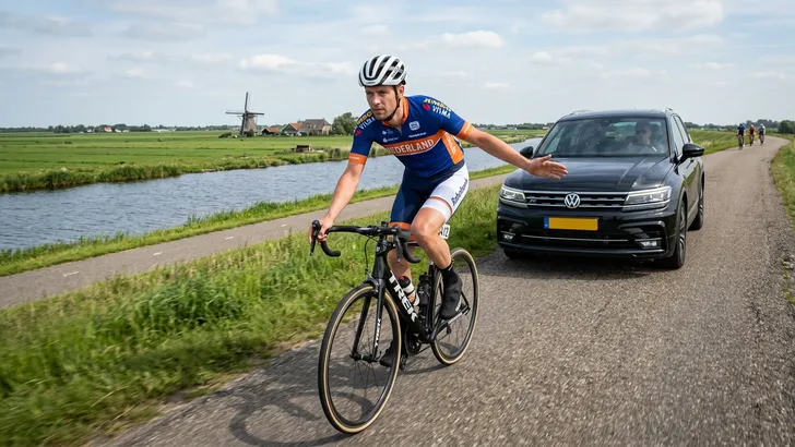 Een wielrenner op een dijk maakt op zijn rug een kalmerend handgebaar naar een donkere SUV die dicht achter hem rijdt.