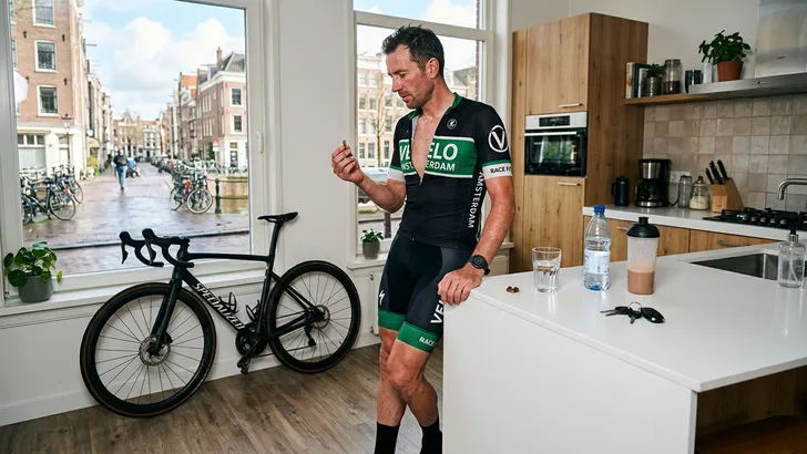 Mannelijke wielrenner zonder helm rust uit in de keuken naast zijn racefiets en gebruikt een supplement met groene thee-extract voor spierherstel.