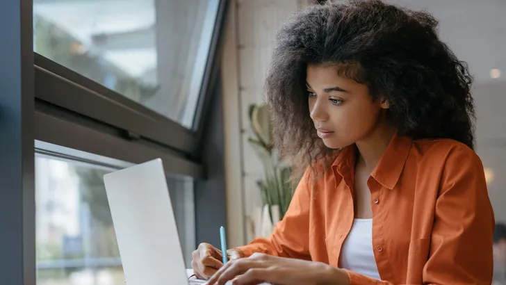 Student using laptop computer, studying, learning language, exam preparation. Pensive African American woman freelancer writes notes working at home