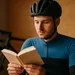 Wielrenner in fietskleding leest een onherkenbaar boek binnenshuis, met een onscherpe racefiets op de achtergrond.