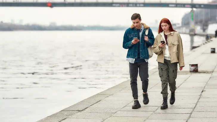 Full length shot of two teenagers totally absorbed in their smartphones, ignoring each other while walking along the riverside together