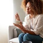 Young relaxed pensive African American woman eating healthy breakfast sitting on the couch. Copy space.