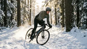 Een wielrenner op een gravelbike neemt een dynamische bocht op een besneeuwd bospad, waarbij sneeuw opspat in het lage zonlicht.