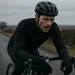 Wielrenner met lange mouwen en handschoenen rijdt uitgeblust over een natte landelijke weg op een grauwe winterdag.