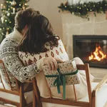 Happy young family in cozy sweaters exchanging stylish christmas gifts on background of fireplace with modern festive mantle and christmas tree with lights. Happy Holidays!