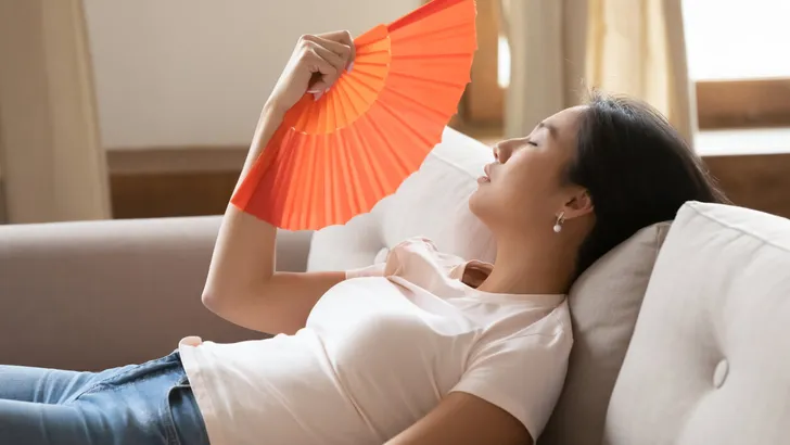 Tired young asian woman using paper fan indoors.