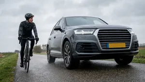 Een wielrenner kijkt bezorgd om terwijl hij op een smalle weg wordt ingehaald door een grote, zwarte SUV die veel ruimte inneemt.