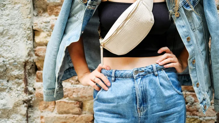 Young beautiful cropped woman standing in street bricks wall, denim jeans clothes, holding belt satchel purse in shoulder, summer fashion trend, close-up details