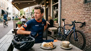Een mannelijke wielrenner van rond de 40 zit zonder helm op een terras met een cappuccino en appeltaart, maar kijkt uiterst zenuwachtig en paranoïde over zijn schouder naar zijn dure carbon racefiets die veilig vlak achter hem tegen een muur staat.