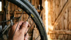 Een close-up van een hand die lucht laat ontsnappen uit het Presta-ventiel van een racefietsband, met een sfeervolle, door warm zonlicht verlichte houten schuur op de achtergrond.
