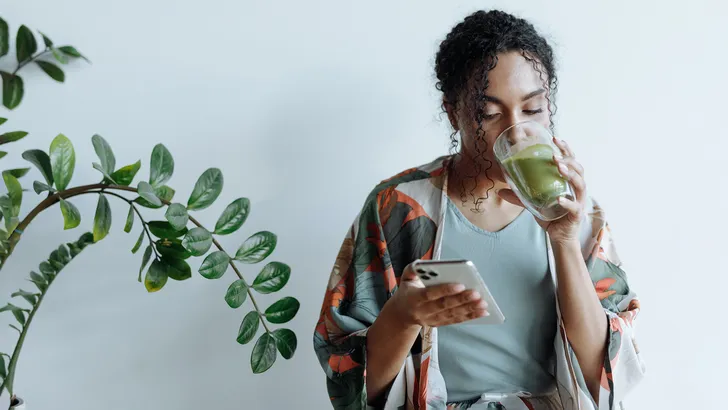 Afbeelding: Vrouw drinkt groene smoothie met telefoon in haar hand