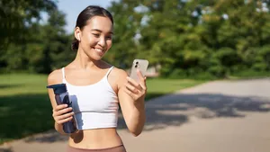 Fitness woman with water bottle and smartphone, jogging in park and smiling, looking at her mobile phone app, checking sport application