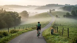 Een mannelijke wielrenner, van achteren gezien, fietst eenzaam een smalle, slingerende heuvelweg op in een glooiend, groen landschap onder een sfeervolle lucht. De afbeelding straalt rust en de inspanning van het klimmen uit.