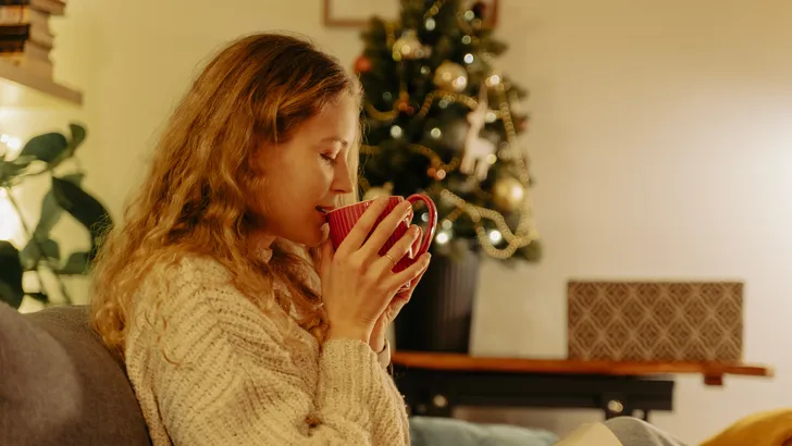 Een vermoeide vrouw zit ontspannen op een bank in een licht interieur met een kop thee, illustratie bij het artikel over moe zijn na de feestdagen en herstel van energie.