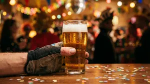 Een close-up van een hand in een wielerhandschoen die een glas bier vasthoudt, met op de achtergrond een sfeervolle, onscherpe carnavalsomgeving met confetti en kleurrijke feestverlichting.