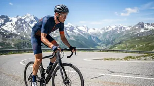 Een zwoegende mannelijke wielrenner met helm 'harkt' staand op de pedalen een steile, zonnige bergpas in de Franse Alpen omhoog, met dramatische toppen en een strakblauwe lucht op de achtergrond.