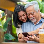 Happy Asian woman teaching elderly father using mobile phone application during having lunch together at restaurant on summer vacation. Family relationship older people mental health care concept.