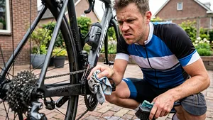Mannelijke wielrenner zonder helm kijkt geërgerd in een Nederlandse straat terwijl hij zijn racefiets poetst, omdat hij met een doekje niet bij de vuile plekken rond de trapas kan komen.